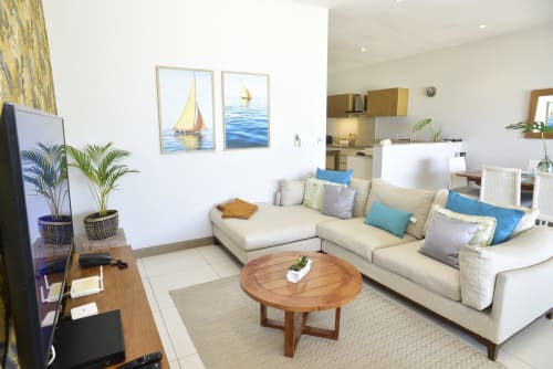 Modern beachfront apartment living room with cream sectional sofa, wooden coffee table, TV, sailboat artwork, and open