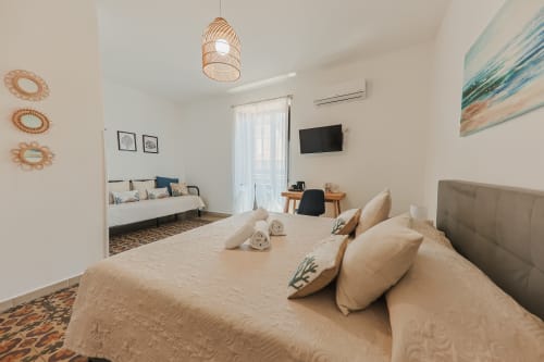 Chambre lumineuse au Casanova B&B à Cefalù avec literie beige, luminaire suspendu tressé et vue sur la ville par les portes du balcon