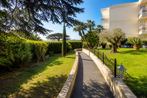 Elegant garden pathway with lush landscaping, olive trees, and white apartment building in Cannes Californie