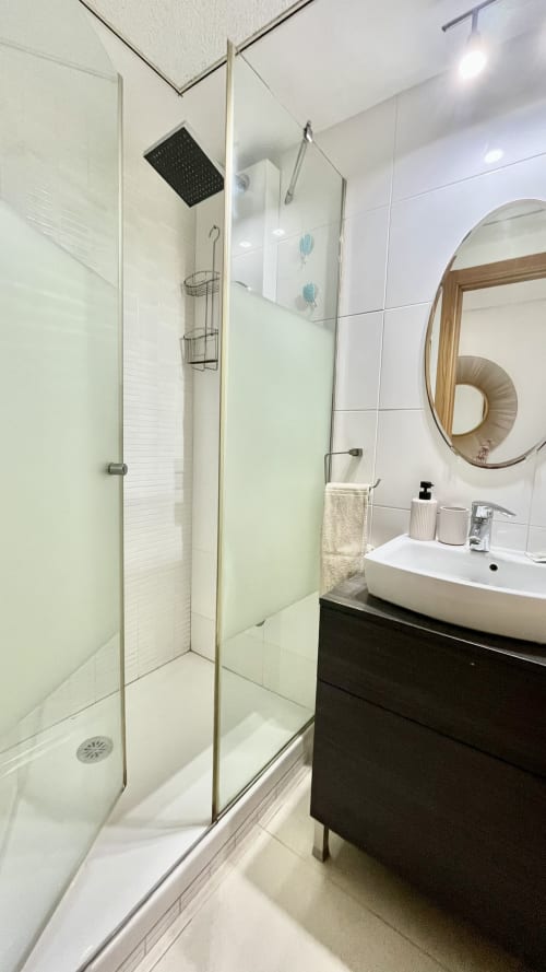 Baño moderno con ducha de cristal, azulejos blancos, lavabo negro y espejo redondo de madera en Pataconablue Valencia