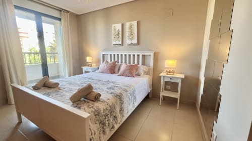 Dormitorio moderno y luminoso en apartamento frente al mar en Valencia, cama blanca, almohadas rosas, acceso a balcón y dos mesitas de noche