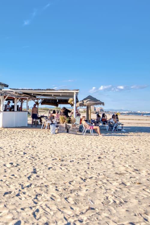 Configuración de bar de playa en Pataconablue Rentals Valencia con huéspedes relajándose bajo sombrillas y pérgola de madera en la playa de arena