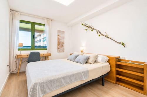 Dormitorio luminoso con cama de madera, escritorio y ventana de marco verde con vistas al mar en un apartamento frente al mar en Valencia