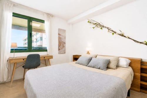 Dormitorio luminoso de apartamento frente al mar en Valencia con vistas al mar, cama de madera, escritorio y ventanas con marco verde