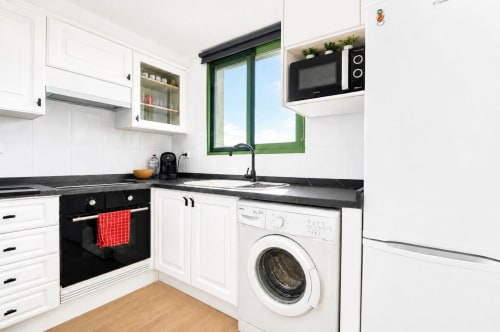 Cocina moderna en apartamento de Valencia con armarios blancos, encimeras negras, lavadora, horno negro y marco verde