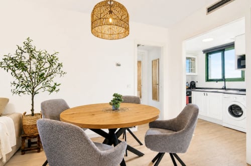 Zona de comedor de apartamento moderno en Valencia con mesa redonda de madera, sillas grises, lámpara colgante tejida, cocina abierta y vistas al mar
