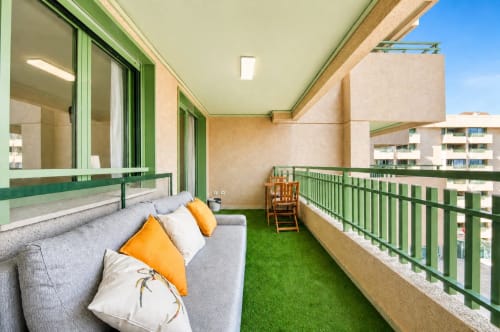 Balcón de apartamento moderno en Valencia con sofá gris, cojines naranjas, césped artificial, vista al mar y barandillas verdes