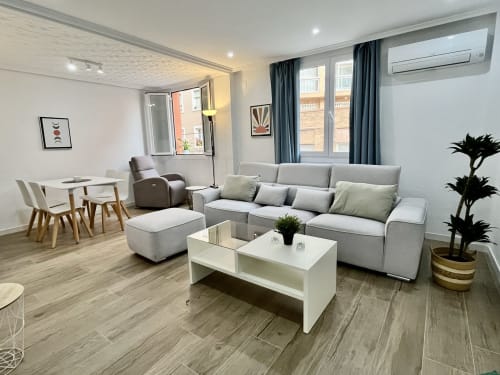 Salón moderno de apartamento en Valencia con sofá gris, mesa de centro blanca, comedor y vistas a la ciudad tras cortinas oscuras