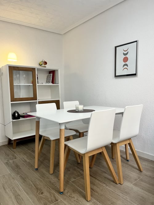 Comedor moderno en apartamento de Valencia con mesa blanca, sillas de madera, mueble de almacenamiento y arte mural de fases lunares