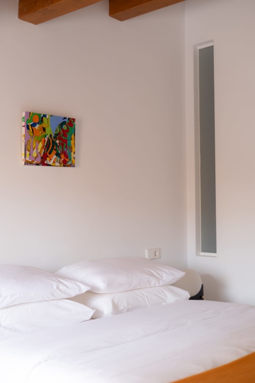 Bright hotel room at Hotel Fragile in Menorca with white pillows, colorful abstract artwork, wooden beams, and minimalist