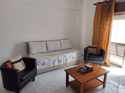 Sala de estar luminosa no apartamento Casa Flor, Albufeira com poltronas pretas, sofá, mesa de madeira e varanda com vista para a cidade