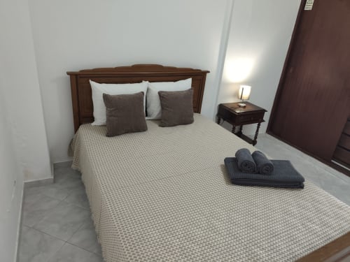 Quarto no Albufeira Inn com cama de madeira, roupa de cama branca, almofadas escuras, criado-mudo com luminária e toalhas enroladas no centro da cidade