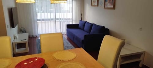 Sala luminosa do apartamento Albufeira Inn com cadeiras de jantar amarelas, mesa laranja, sofá azul e kitchenette moderna