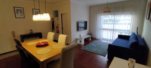 Área de estar integrada no apartamento de Albufeira com mesa de jantar, cadeiras amarelas, sofá azul, TV e janelas iluminadas