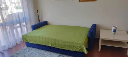 Quarto luminoso no Albufeira Inn com roupa de cama verde-lima no sofá-cama azul, criado branco e luz natural da varanda