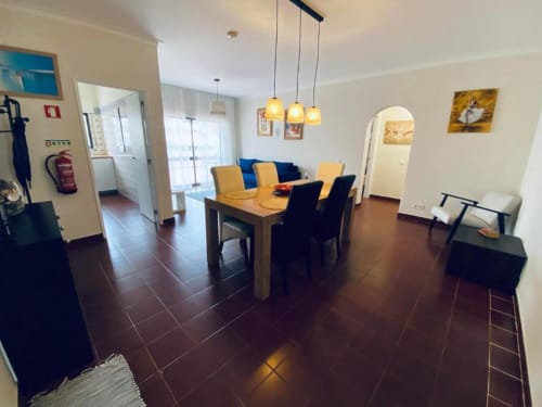 Sala de estar e jantar do Albufeira Inn com mesa de madeira, cadeiras amarelas, sofá azul e luminárias pendentes em Praia dos
