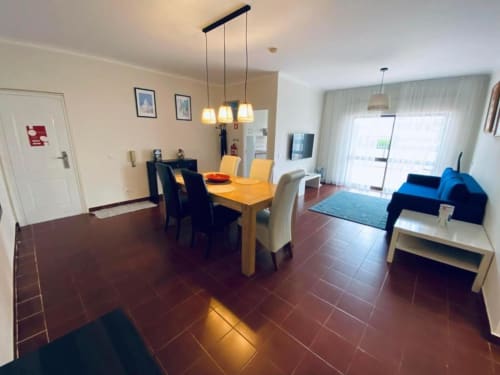 Sala de estar e jantar do Albufeira Inn com mesa de madeira, cozinha e sofá azul sobre piso de azulejo terracota