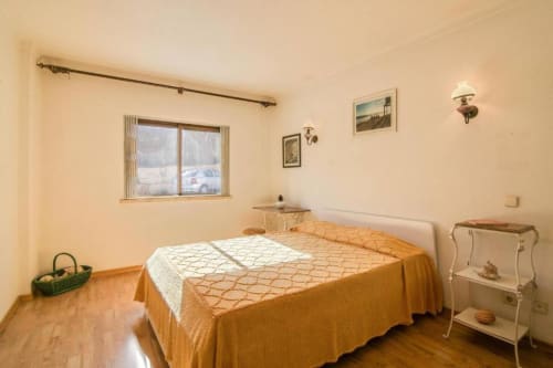 Quarto luminoso no Albufeira Inn com cama de colcha laranja, vista da janela, arandelas na parede e piso de madeira