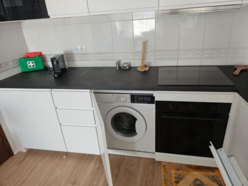 Cozinha moderna em apartamento de Albufeira com bancada preta, armários brancos, máquina de lavar, forno e painel de azulejos
