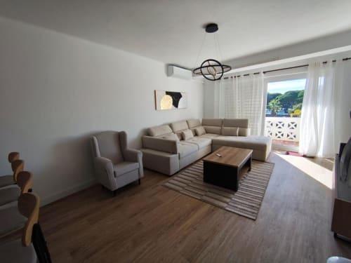 Sala de estar moderna na Albufeira Inn com sofá bege em L, poltrona, mesa de centro, piso de madeira e varanda