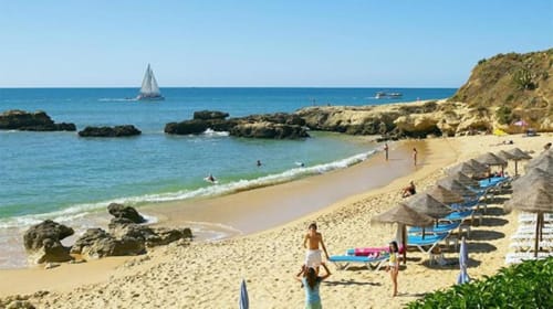 Praia dos Aveiros ensolarada com areia dourada, guarda-sóis azuis, afloramentos rochosos, veleiro e vistas de falésias