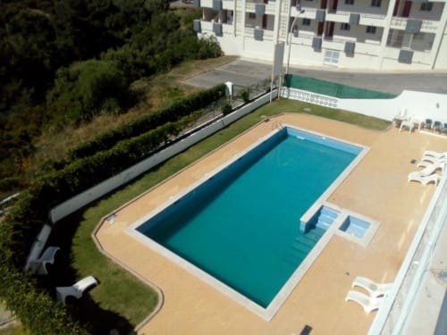 Vista aérea da piscina turquesa do Albufeira Inn cercada por espreguiçadeiras, plataforma arenosa e edifício moderno de apartamentos