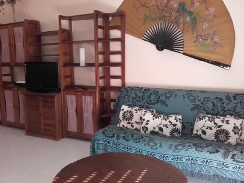 Sala de estar brilhante com sofá azul-petróleo, ventilador decorativo, rack de madeira com TV e almofadas estampadas