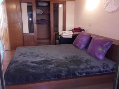 Quarto espaçoso na Albufeira Inn com cama de casal escura, almofadas roxas, guarda-roupa em madeira e paredes rosadas no apartamento T1