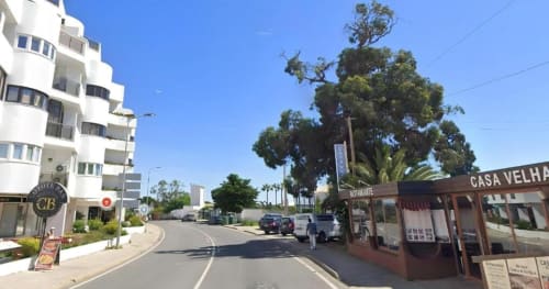 Vista da rua do Albufeira Inn com edifício branco de apartamentos, restaurante e grande árvore sob céu azul