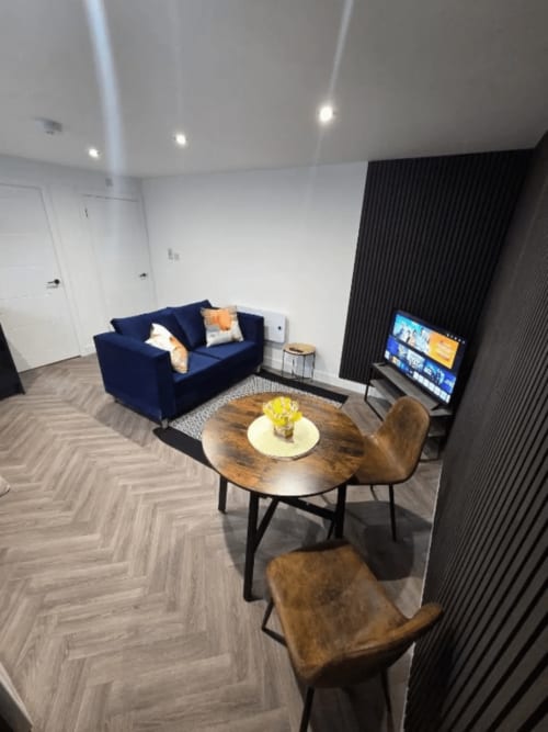 Modern apartment living space with navy sofa, wooden table, brown chairs, TV, and herringbone flooring at JS Apartments