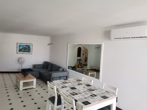 Sala de estar integrada e moderna no Albufeira Inn com mesa de refeições, sofá cinzento, kitchenette e grande espelho no apartamento T1.
