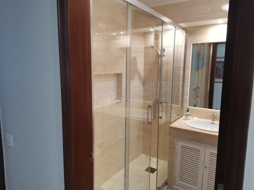 Banheiro moderno com grande boxe de vidro, azulejos bege, bancada branca e detalhes em madeira no Albufeira Inn,
