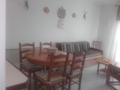 Sala de estar com mesa de jantar e cadeiras de madeira no apartamento Albufeira Inn, paredes brancas com decoração náutica