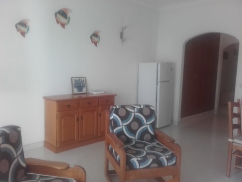 Sala de estar do apartamento Albufeira Inn com móveis de madeira, almofadas estampadas, frigorífico branco e parede decorativa
