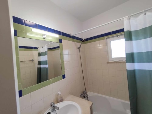 Banheiro moderno com acabamento em azulejos verde e azul, espelho, pia, vaso sanitário e banheira com chuveiro na villa Albufeira Inn