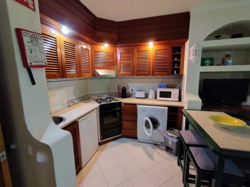 Cozinha moderna no estúdio da Albufeira Inn com armários em madeira, eletrodomésticos brancos e zona de refeições com vista para o jardim.