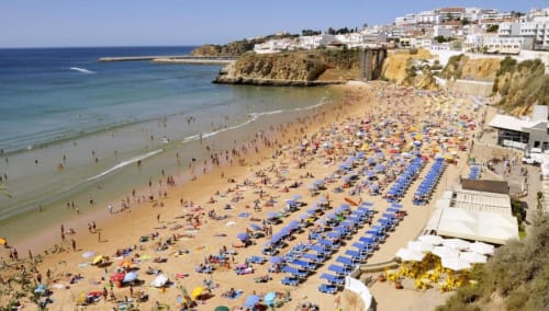 Praia de Albufeira repleta de areia dourada, guarda-sóis azuis, edifícios no topo dos penhascos e águas turquesas tranquilas do Mediterrâneo.