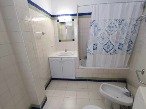 Casa de banho moderna com azulejos brancos, detalhes azuis, espelho, lavatório, e cortina de chuveiro com padrão azul na villa Albufeira Inn