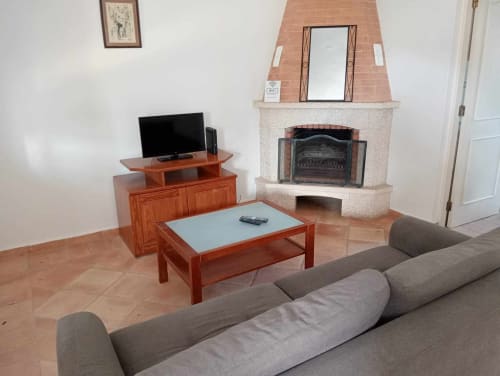 Sala de estar luminosa com sofá cinzento, móveis de madeira, lareira e TV na moradia de Albufeira com piso em azulejo de barro.