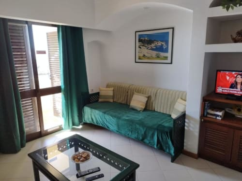 Sala luminosa do estúdio com sofá verde-azulado, TV, kitchenette e vista de jardim no Albufeira Inn, Portugal