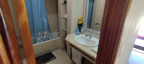 Banheiro moderno com pia branca, espelho, chuveiro com cortina azul e portas de madeira no apartamento Albufeira Inn