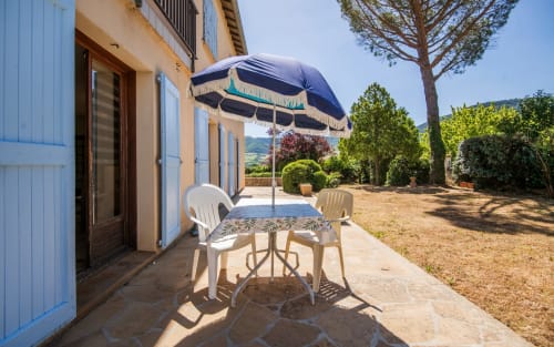 Soleada terraza de villa en Millau con sombrilla azul, sillas blancas, jardín paisajístico con árboles y vistas al campo.