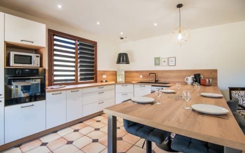 Moderna zona cocina-comedor en villa Millau con muebles blancos, encimeras de madera, persianas a rayas y mesa puesta