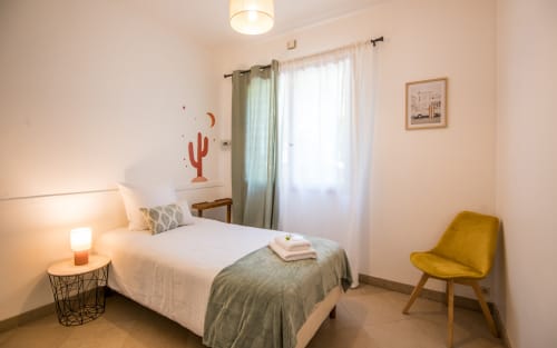 Dormitorio luminoso en villa de Millau con cama blanca, silla amarilla de mediados de siglo, arte mural de cactus y vistas al jardín