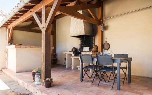 Terraza cubierta de la villa La Conciergerie en Millau con zona de comedor, chimenea y vigas de madera rústica