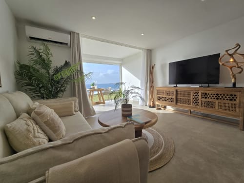 Salon d'appartement vue mer de luxe avec sièges crème, téléviseur écran plat, meuble média en bois, vue océan depuis balcon