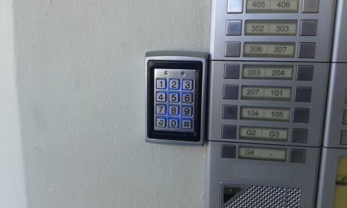 Painel de controle de acesso na entrada do apartamento Albufeira Inn com botões numerados e painel do elevador adjacente do edifício
