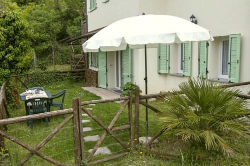 Outdoor garden patio at Casa di Cinti with white umbrella, wooden fence, green shutters, and lounge seating in Loiano