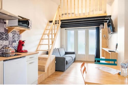 Studio compact à la Villa des Palmiers avec lit mezzanine, kitchenette, canapé gris et grandes fenêtres à Villeneuve-sur-Lot
