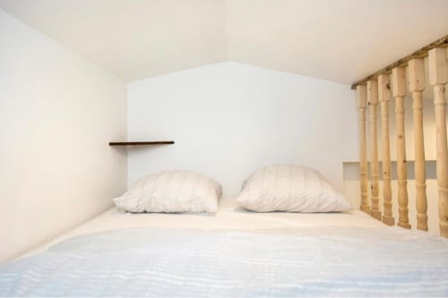 Chambre de studio minimaliste à la Villa des Palmiers avec oreillers blancs, lit bébé en bois, rideaux beige et étagère murale